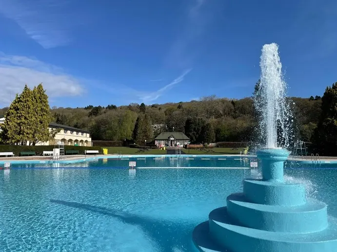 Large outdoor swimming pool with central fountain, surrounded by trees and hillside on a clear sunny day.