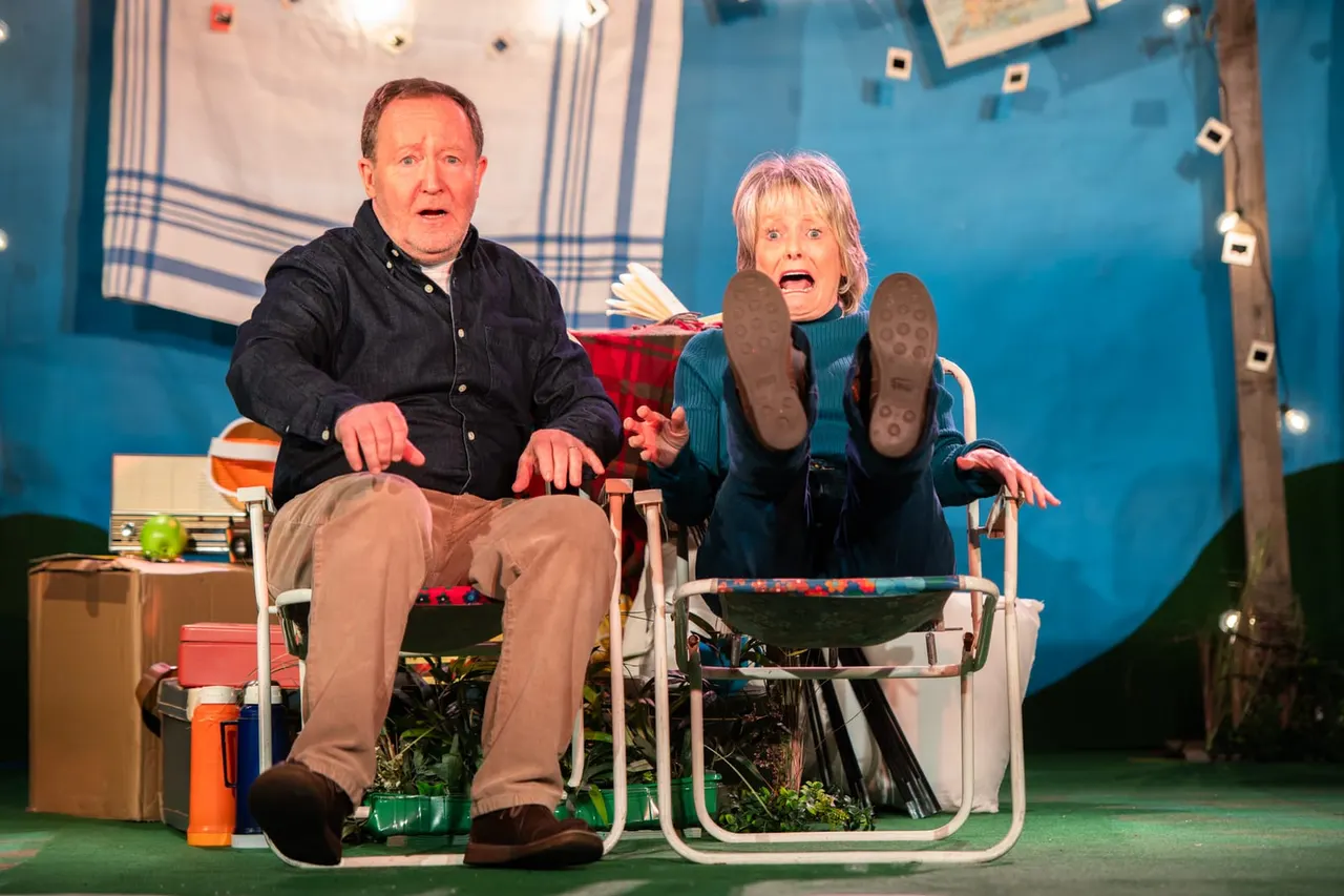 Two actors seated on camping chairs on stage during a comedy performance, with one raising their legs in an exaggerated gesture.