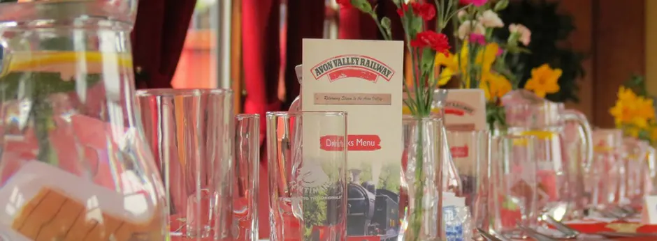 Dining table set with glasses, cutlery, napkins and fresh flower arrangements, with an Avon Valley Railway menu card at centre.