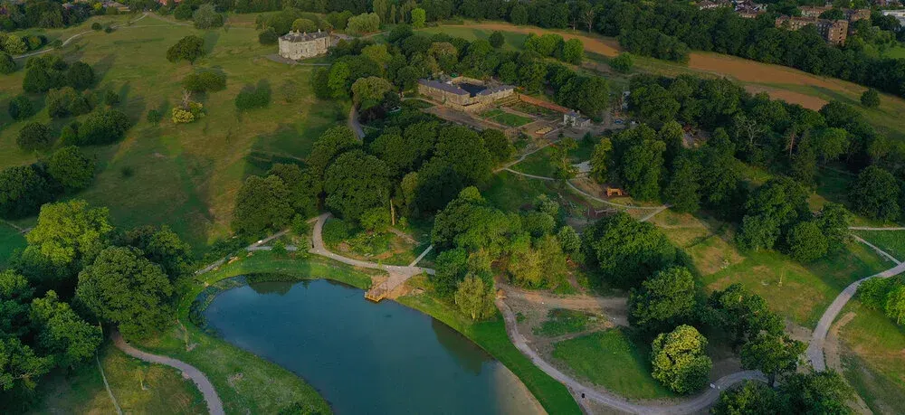 Aerial view of a large pond surrounded by woodland and parkland with pathways and historic buildings visible in the distance.