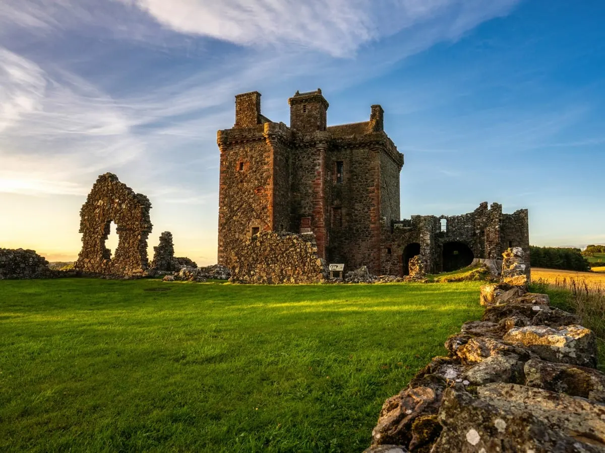 Ruined stone castle with multiple towers standing in a green field under blue sky.