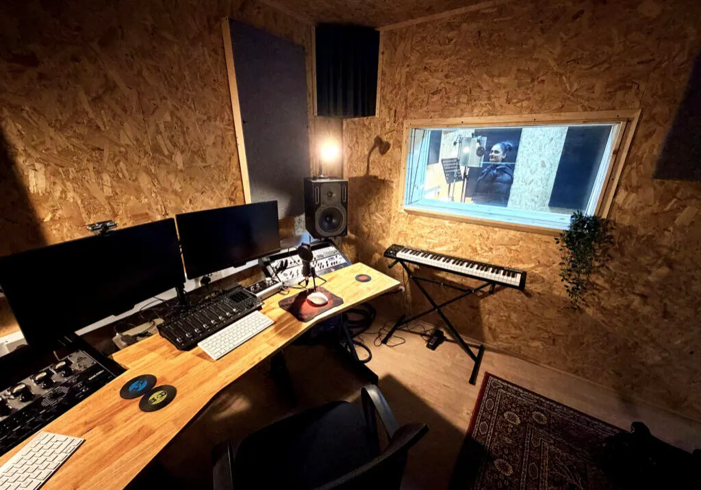 Music production studio with cork-lined walls, workstation featuring monitors, keyboards, and audio equipment.
