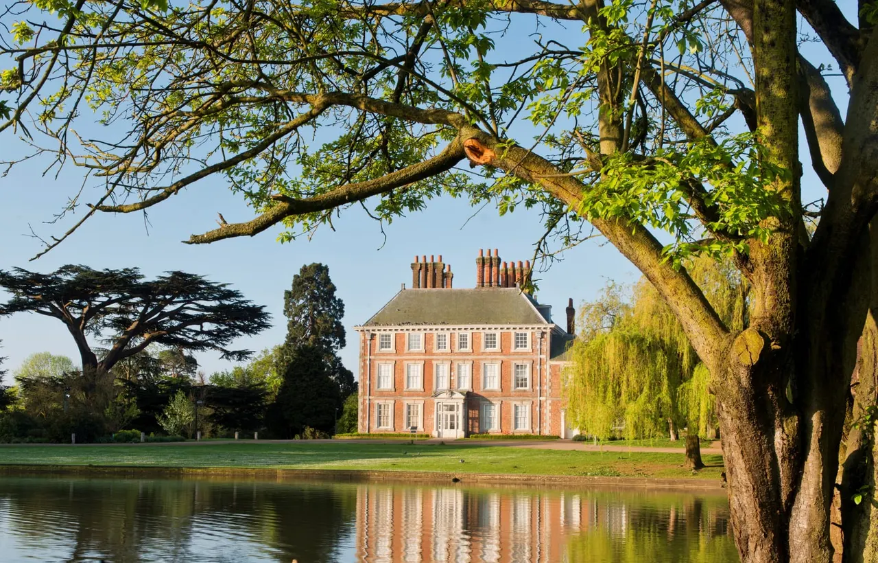 A Georgian brick mansion viewed across a lake, framed by mature trees with spring foliage.