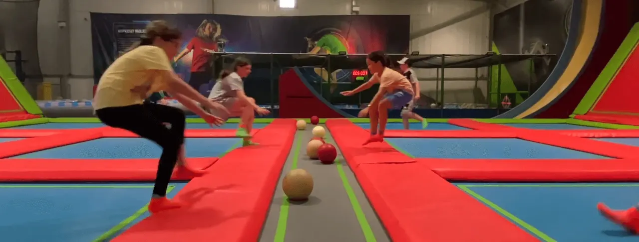 Adults and children playing dodgeball on colourful padded trampolines in an indoor trampoline park.