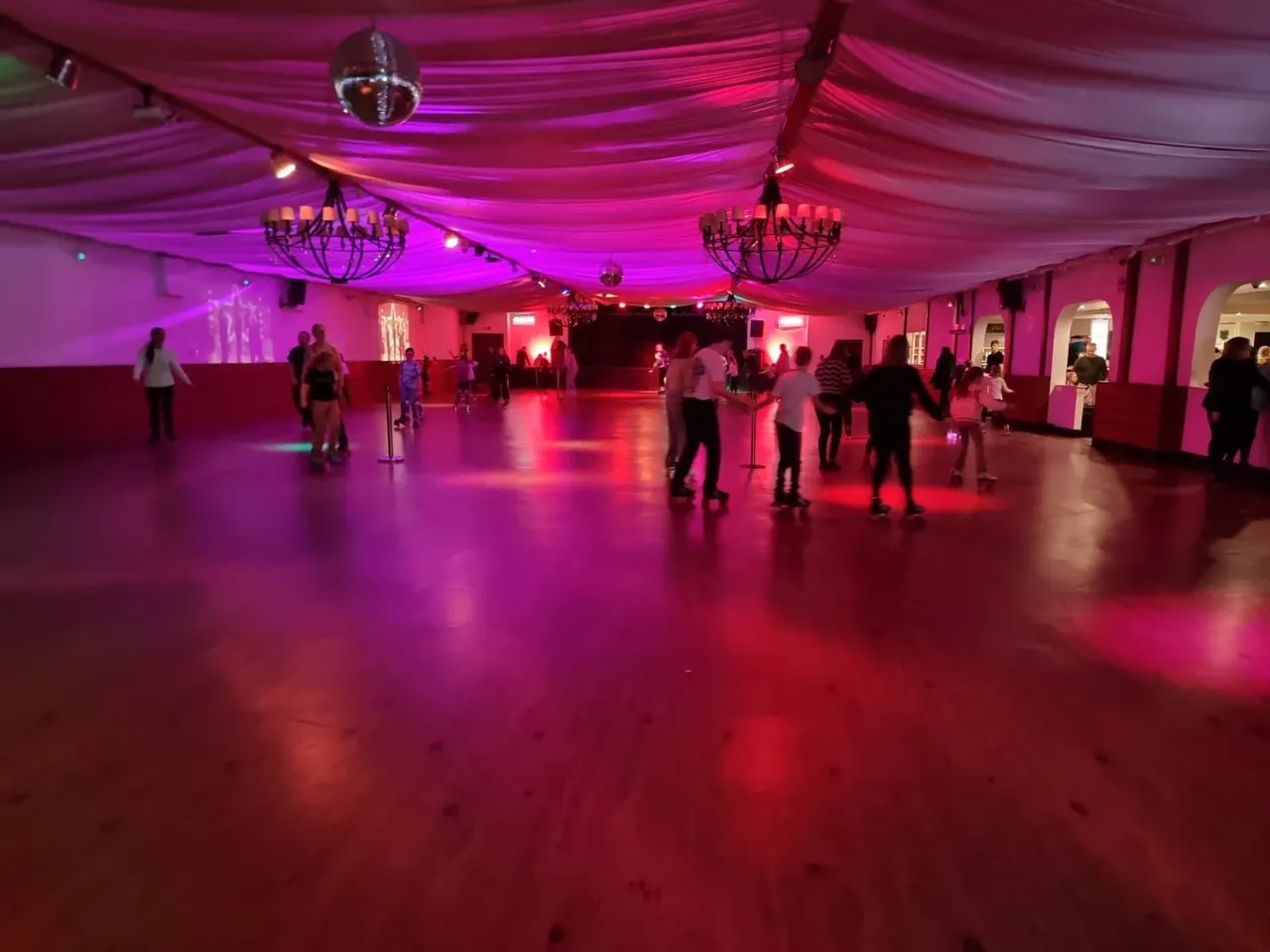 Roller skating venue with visitors skating on a polished floor under vibrant magenta and pink lighting, with draped fabric ceiling and chandeliers.