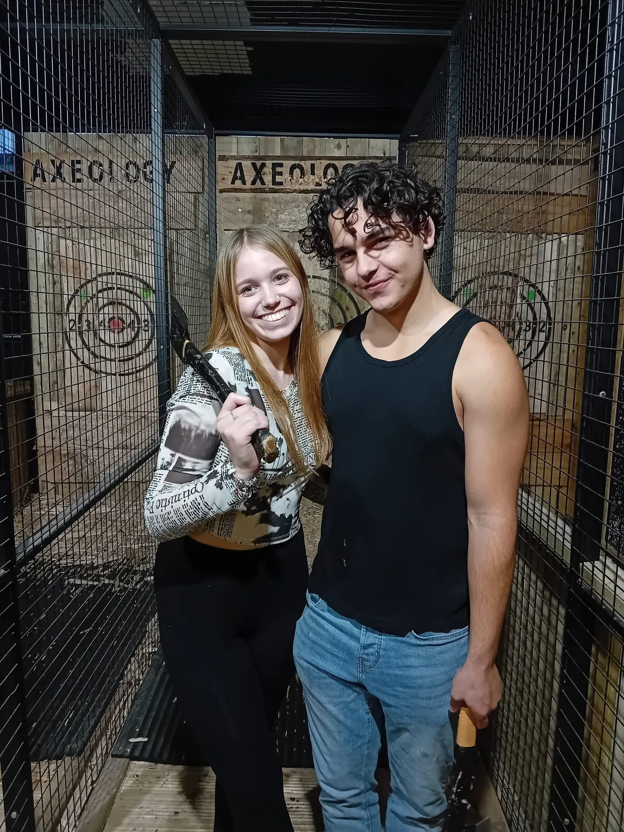 Two young people posing together inside a metal cage at an axe-throwing venue.