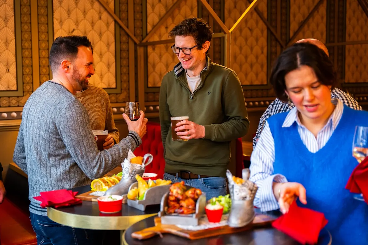 Three people socialise whilst holding drinks and eating from a buffet table in an ornate gold-panelled interior.