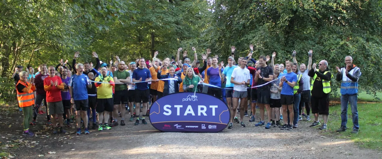Large group of people gathered at a running event start line in a tree-lined forest path, with arms raised.