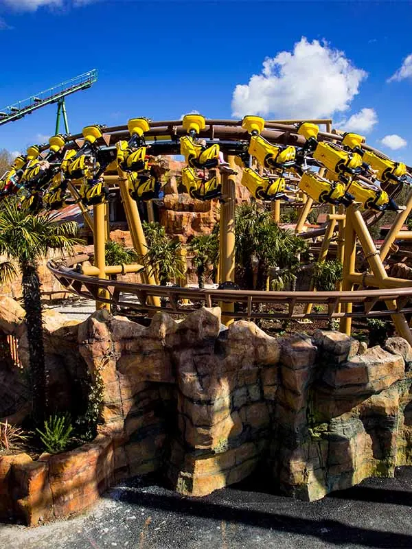 Yellow roller coaster with riders soaring over rocky landscape and tropical plants under blue sky.