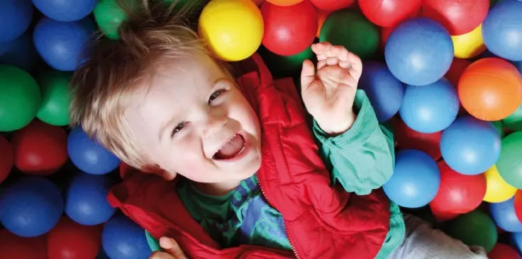 Young child laughing and playing in a colourful ball pit with red, blue, green and yellow balls.