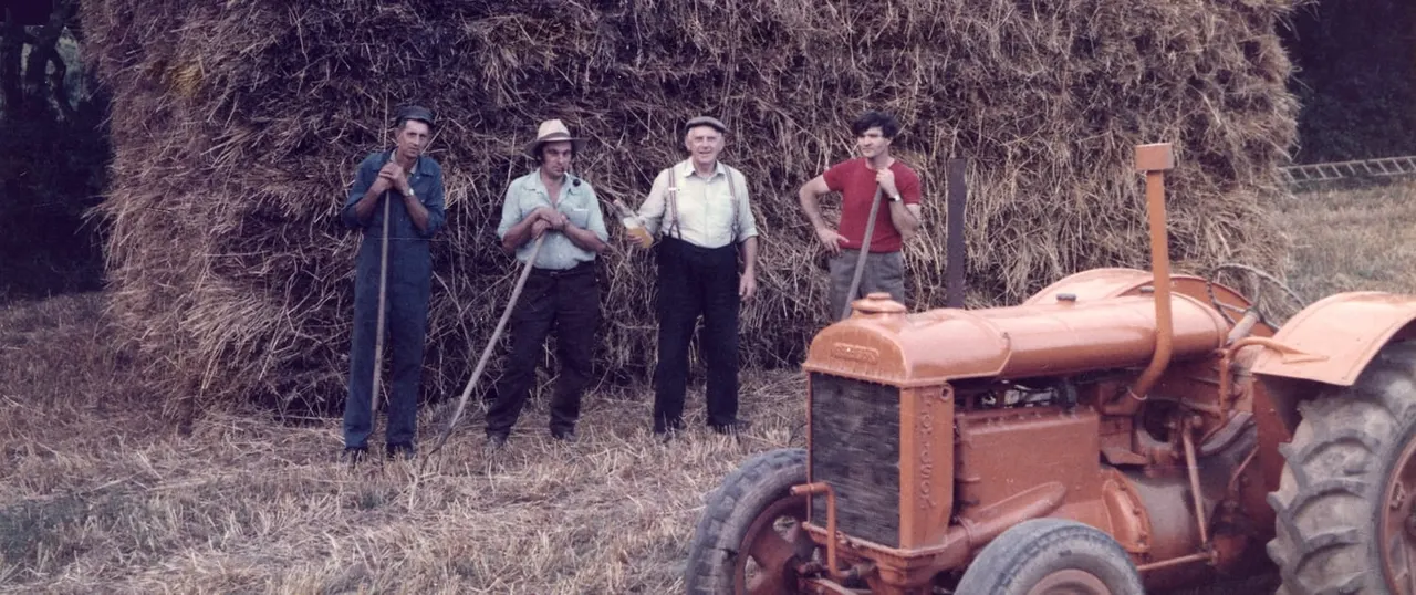 Four men with farming tools stand beside a vintage tractor in a rural setting with dried vegetation.