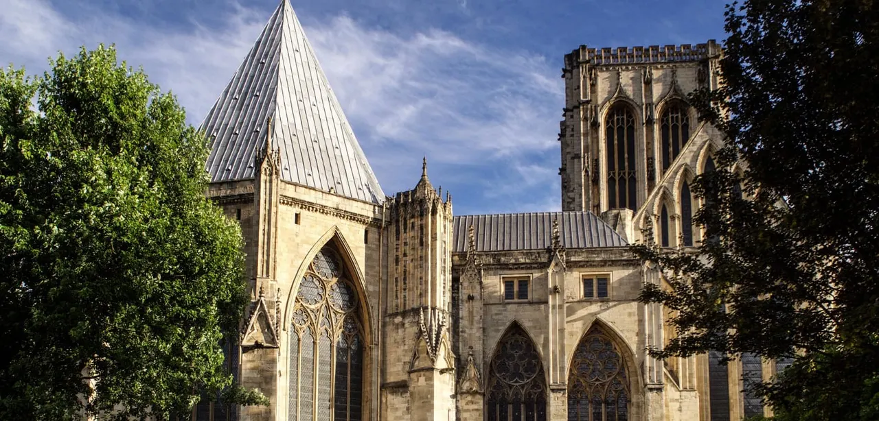 Gothic cathedral with stone walls, pointed arch windows, and metal roof, framed by green foliage under blue sky.