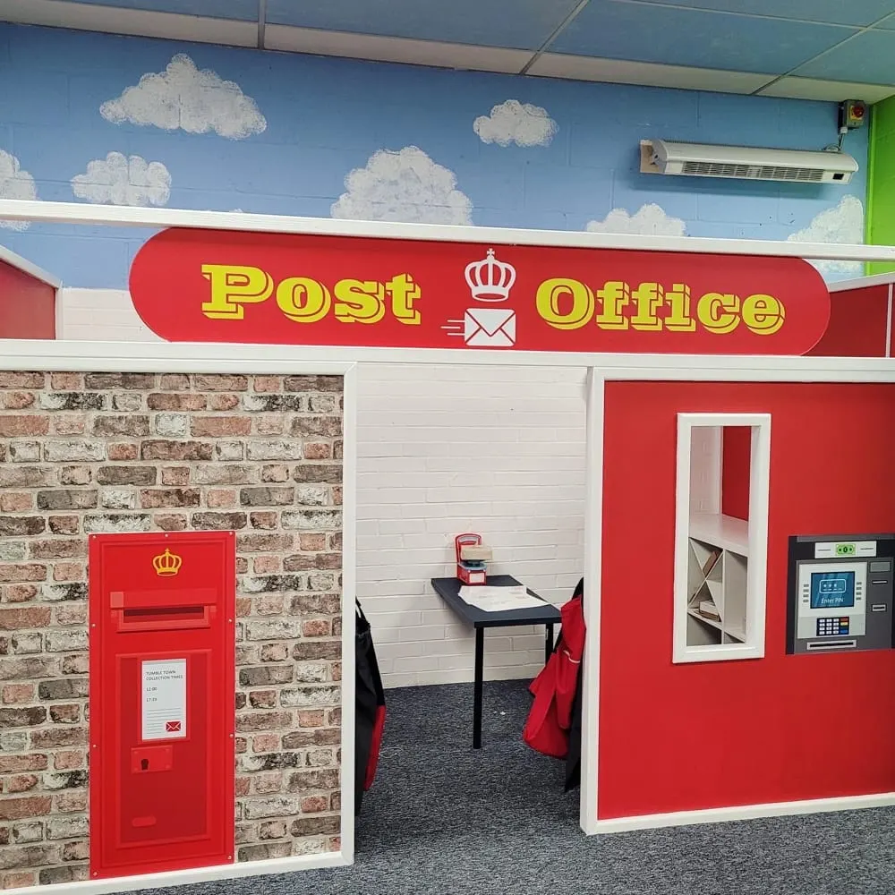 A child-sized post office playhouse with red and white panels, a red post box, counter with till and mail sorting desk inside.