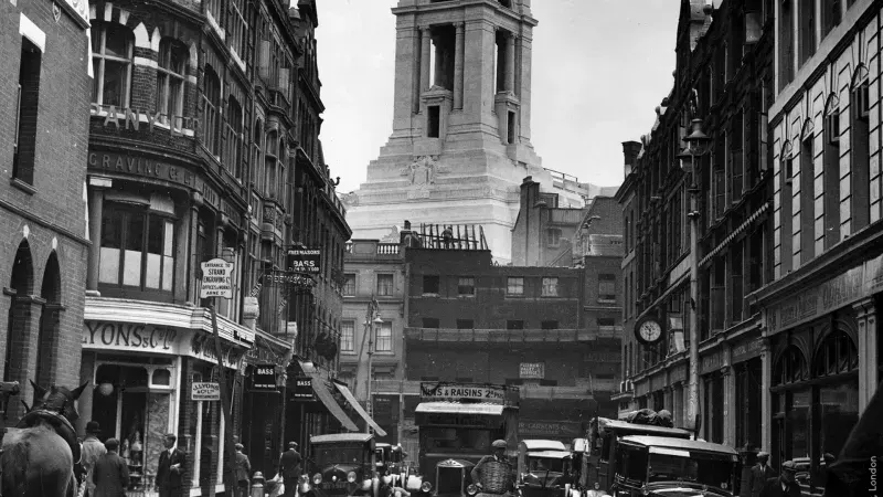 Historic street scene with a church tower visible beyond Victorian buildings and early motor vehicles.