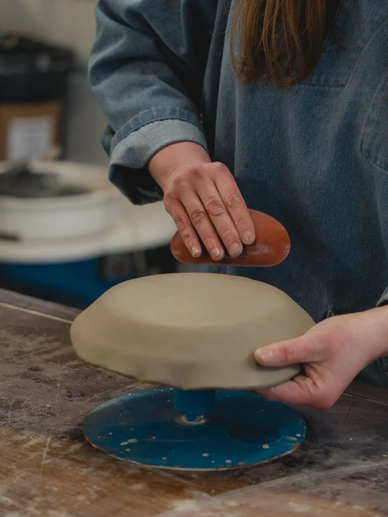 Person in denim shirt hand-finishing a ceramic plate with a wooden tool at a pottery workshop.
