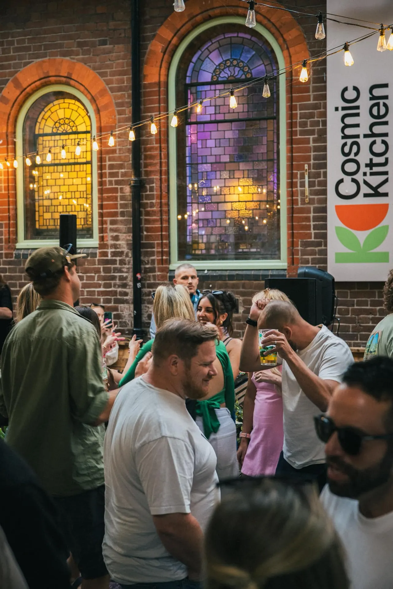Group of people socialising indoors at a brick venue with illuminated arched windows and hanging lights.