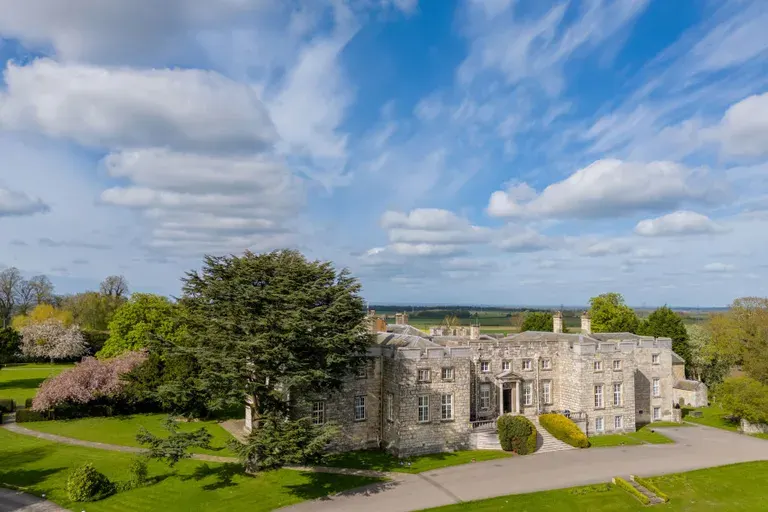 Stone castle with manicured gardens and tree-lined grounds under a blue sky with white clouds.
