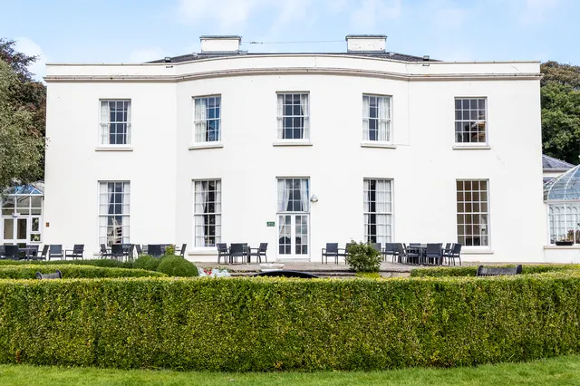 White Georgian mansion with seating area in front, bordered by manicured hedge.