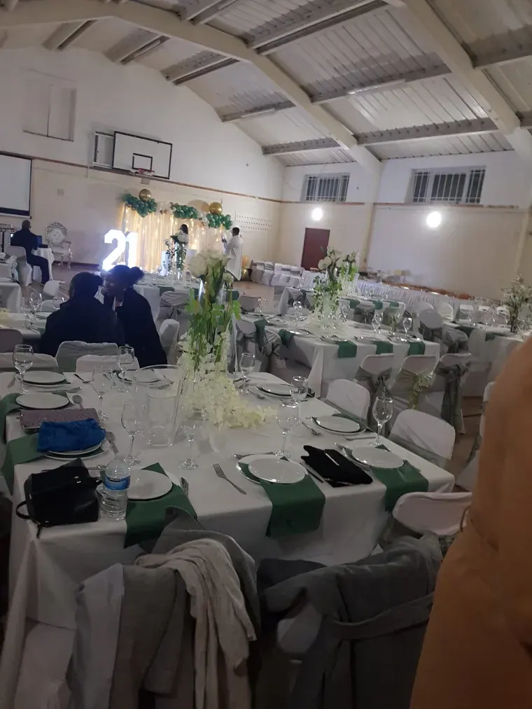 Wedding reception hall with long tables set with white linens, green napkins, floral centrepieces, and place settings, guests seated and mingling.