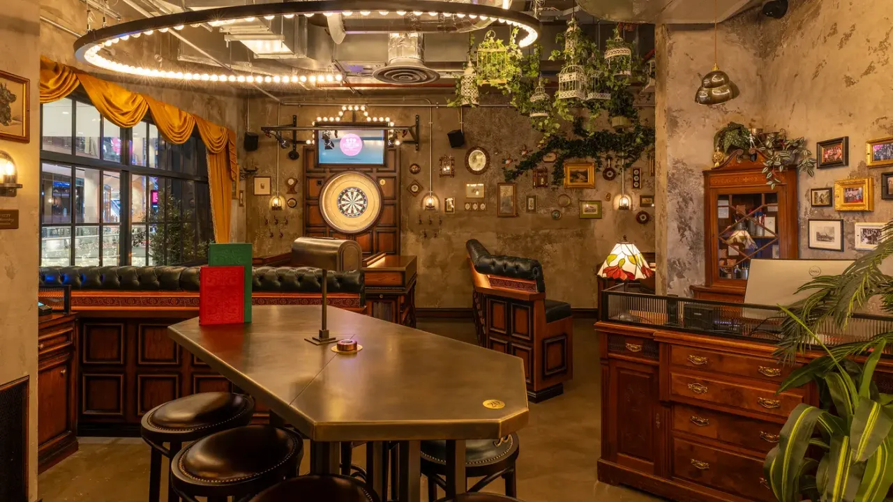 Vintage-style indoor games room with darts boards, leather seating, wooden furniture, decorative clocks and hanging plants under warm lighting.