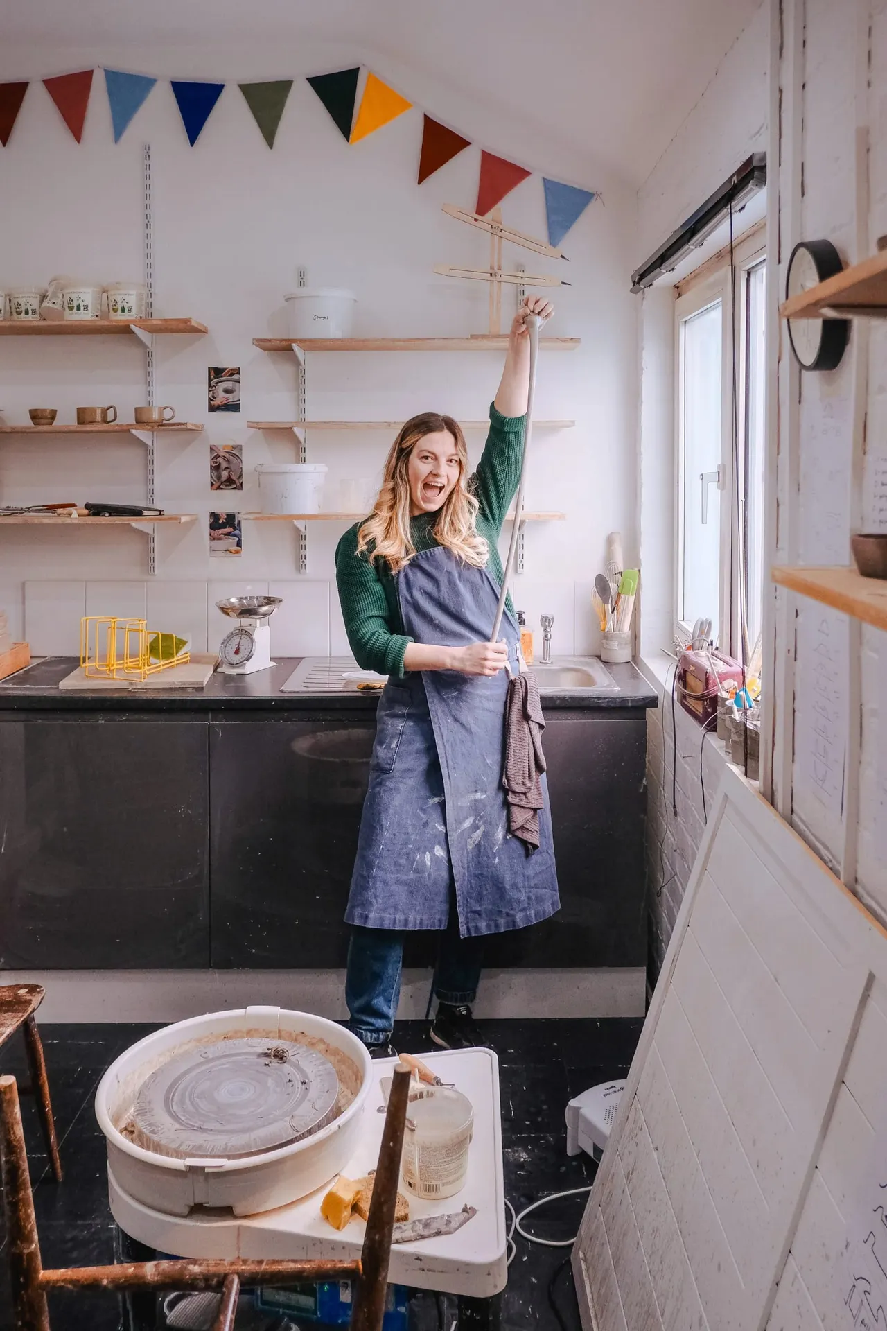 A woman in a denim apron stands cheerfully in a bright pottery studio with a pottery wheel in the foreground and wooden shelves displaying ceramics and craft supplies.