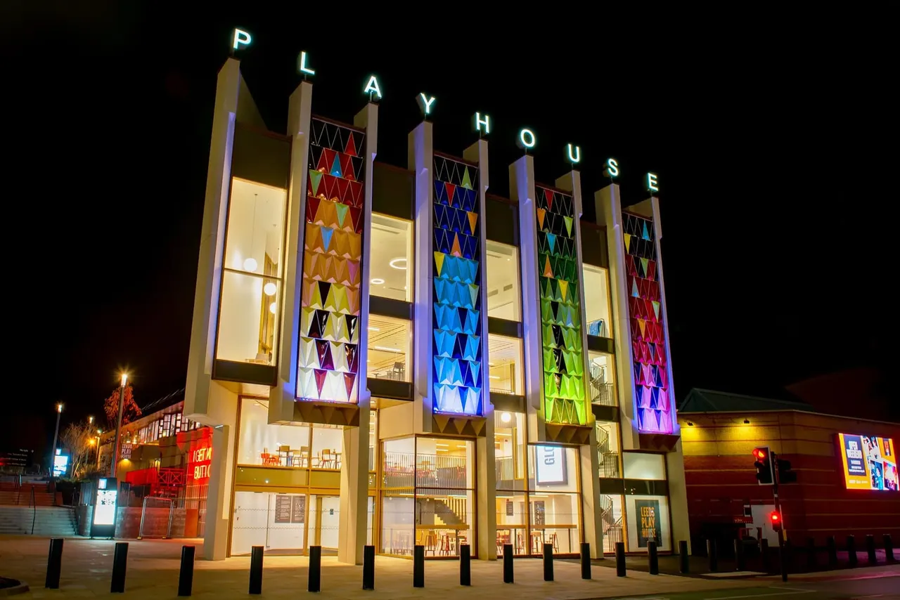 Nighttime exterior of a modern theatre building with tall illuminated columns spelling "PLAYHOUSE" in neon lettering.