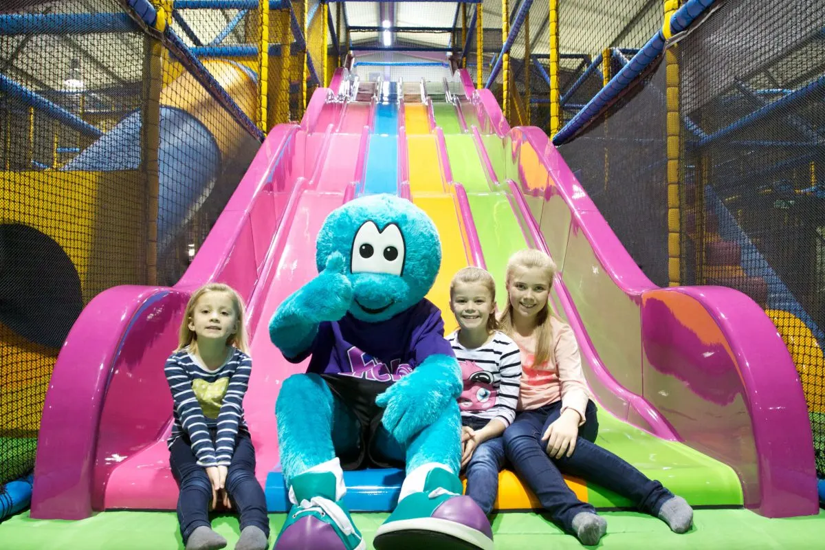 Three children and a blue costumed character sitting together at the bottom of a colourful indoor slide in a play centre.
