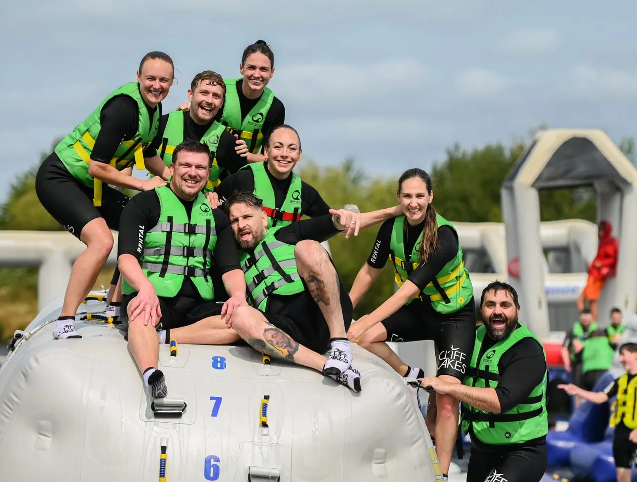A group of eight people wearing bright green and black wetsuits pose together on an inflatable white water sports raft, smiling at the camera.