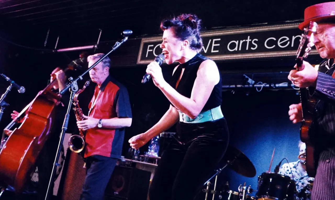 Band performing on stage with string instruments and microphones under pink and blue stage lighting.