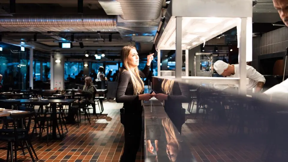 A woman stands at a counter in an evening-lit restaurant or bar with dining tables and other guests visible in the background.
