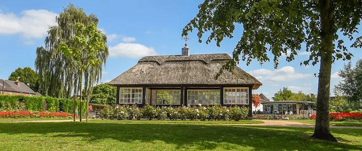 Traditional thatched cottage with manicured lawns, mature trees, and flower borders in a park setting.