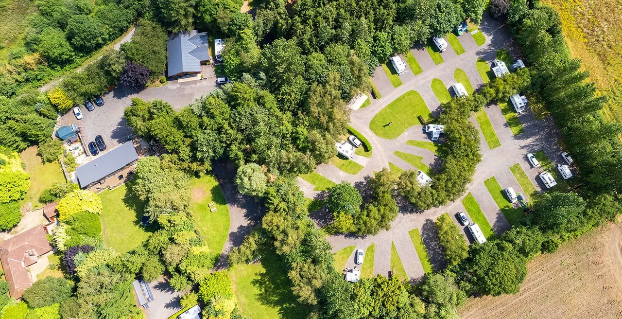 Aerial view of a caravan park nestled amongst trees, with pitches, vehicles, facilities buildings and green spaces.