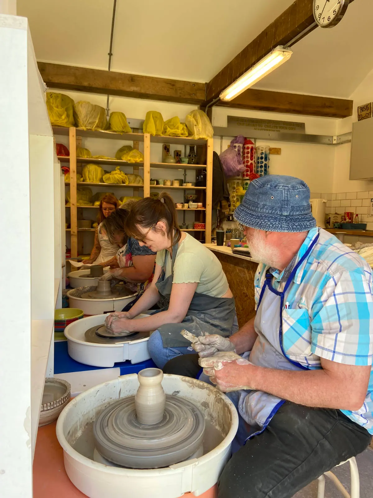 A man and woman work at pottery wheels in a studio, shaping clay whilst another person watches in the background near shelves of finished ceramic pieces.
