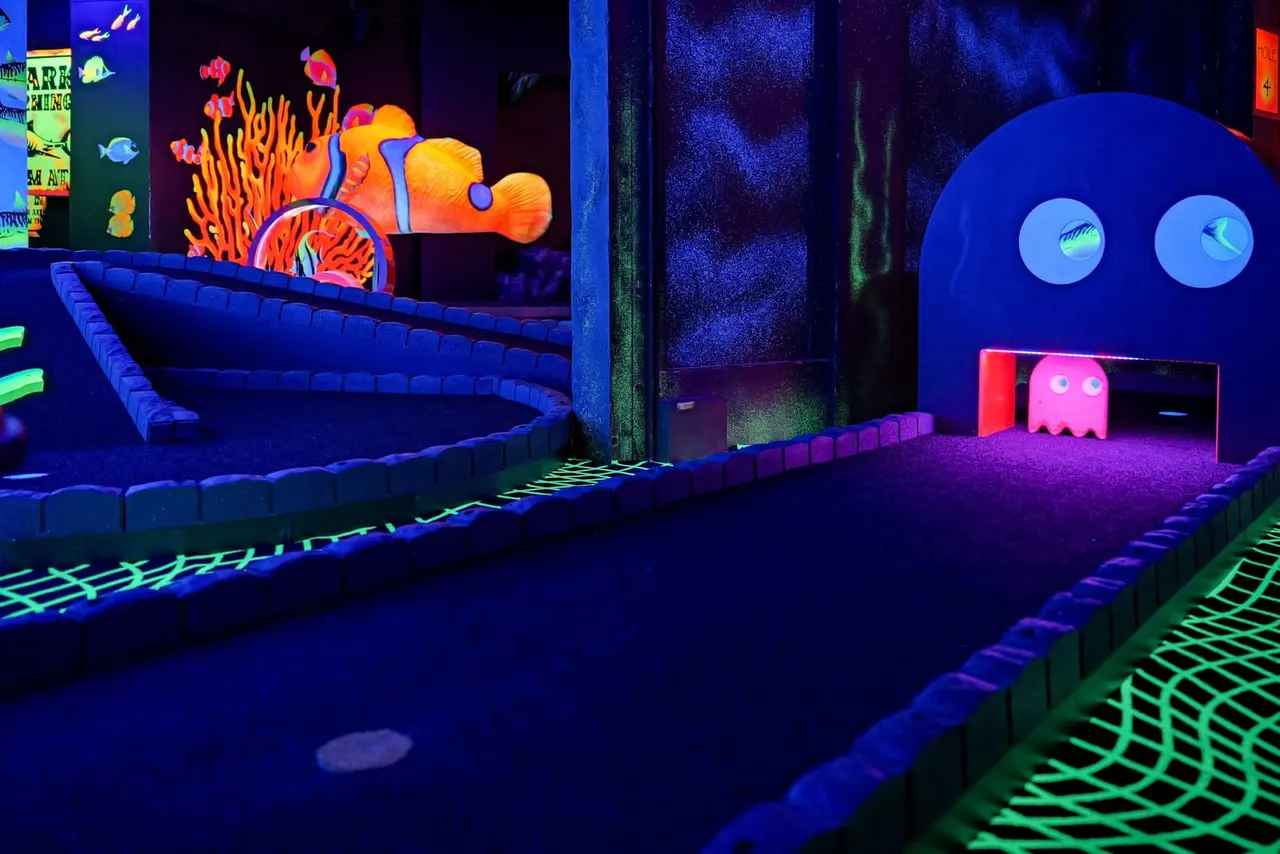 Neon-lit indoor mini golf course with glow-in-the-dark obstacles, including coral reef and ghost-themed designs under ultraviolet lighting.