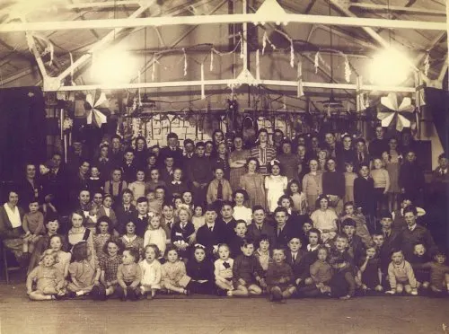 Large group of people posed together indoors in a hangar-like space with exposed beam ceiling and large fan visible.