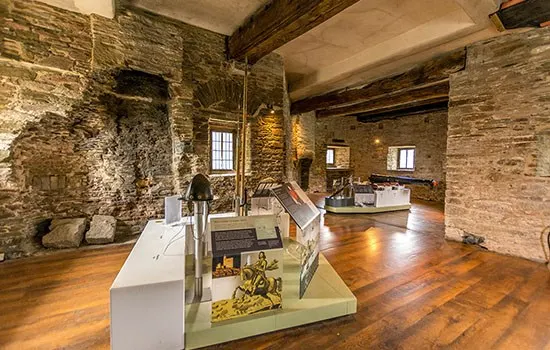 Interior of a stone-walled museum gallery with wooden flooring and display cases showing artefacts.