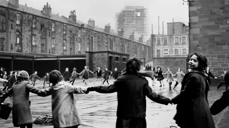 Children playing and forming a circle in a wet urban courtyard surrounded by tenement buildings.