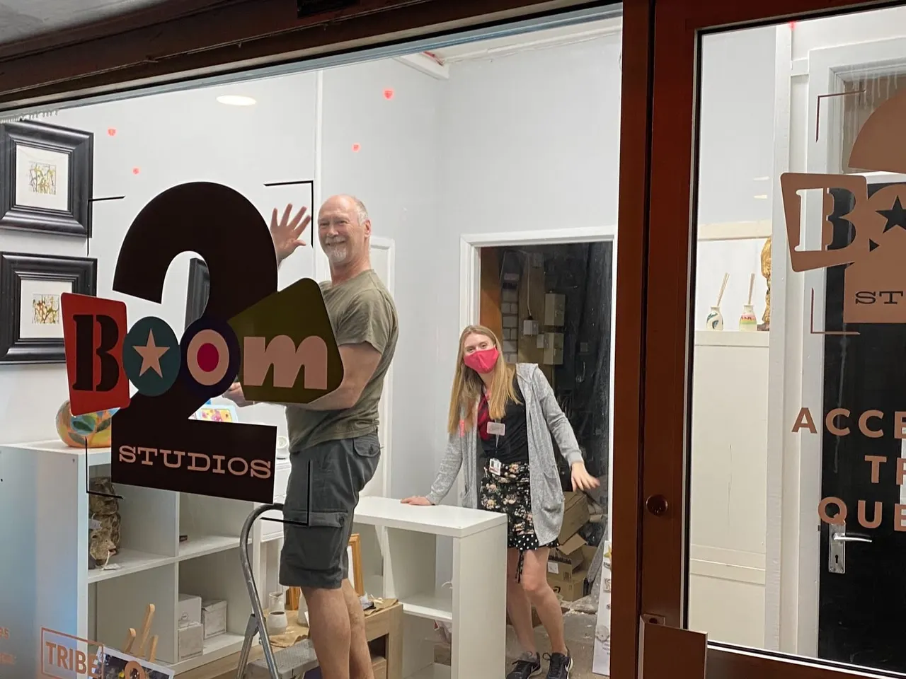 A man and woman stand inside an art studio reception area, with the woman wearing a pink face mask at a white desk.