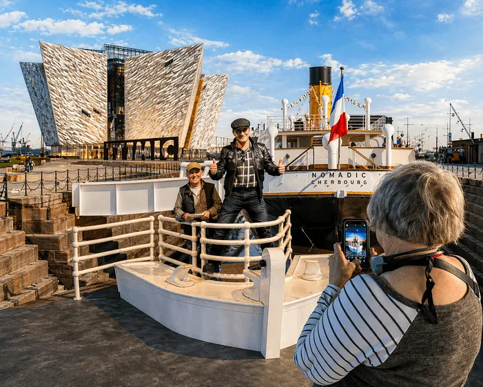 Visitor photographing two people posing in a white boat prop at Belfast waterfront with Titanic Museum and moored ship behind.