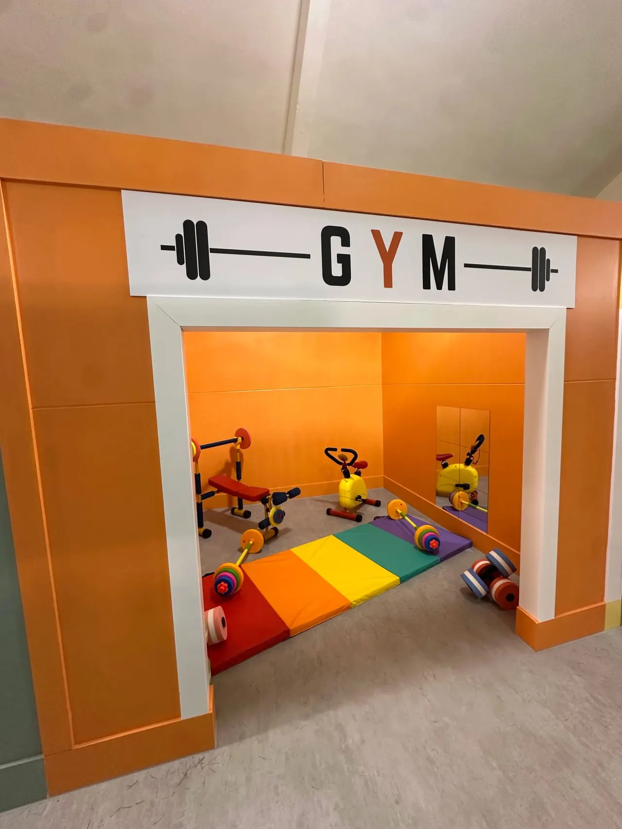 Children's gym play area with colourful exercise equipment and foam mat, viewed through orange-framed entrance.