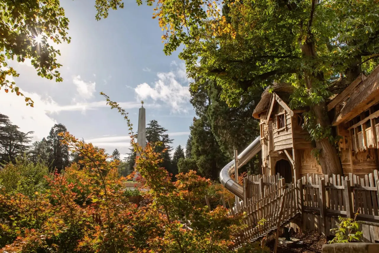 Wooden children's play structure with slide nestled among autumn trees, with a monument visible in the distance.