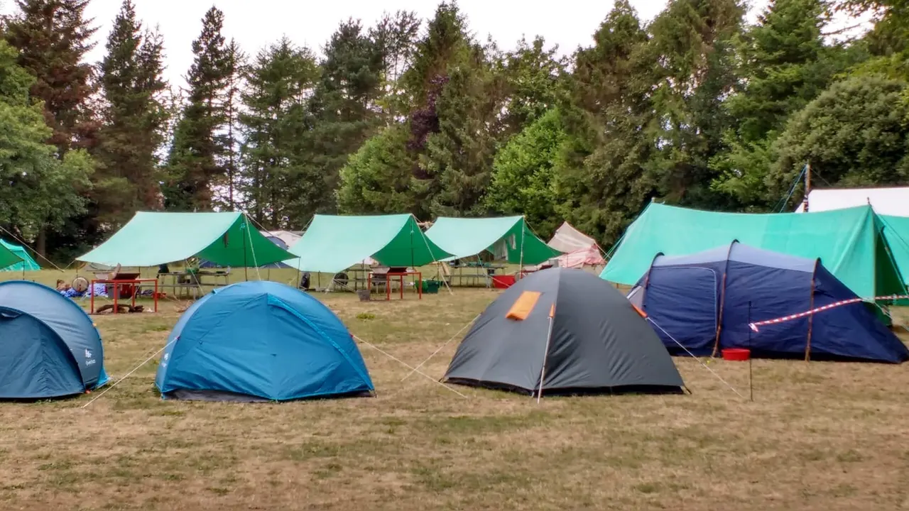 Camping field with multiple blue and grey dome tents, turquoise canopies, and conifer trees in background.