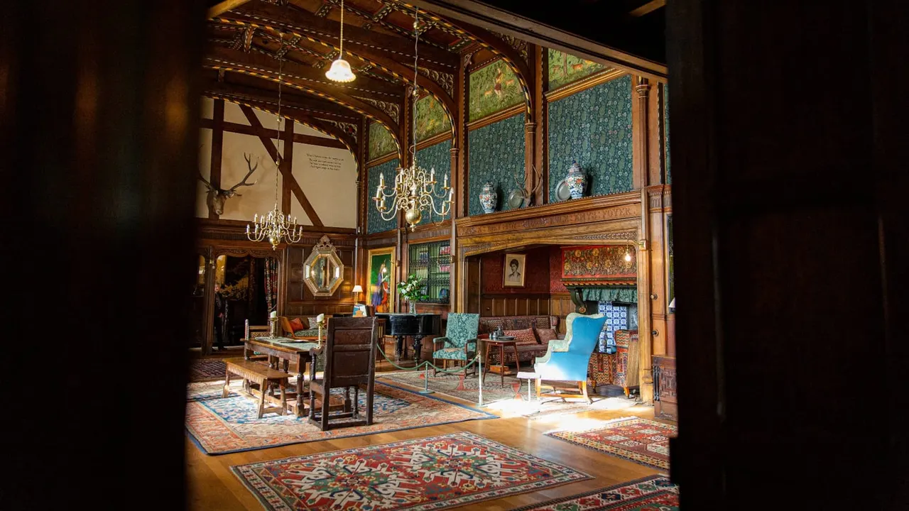 Historic manor interior with ornate wooden panelling, decorative wallpaper, period furniture, chandeliers and artwork displayed throughout the room.