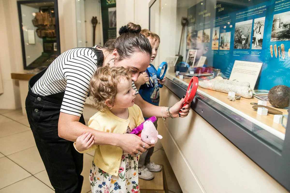 An adult and young children using magnifying glasses to examine artefacts in a museum display case.