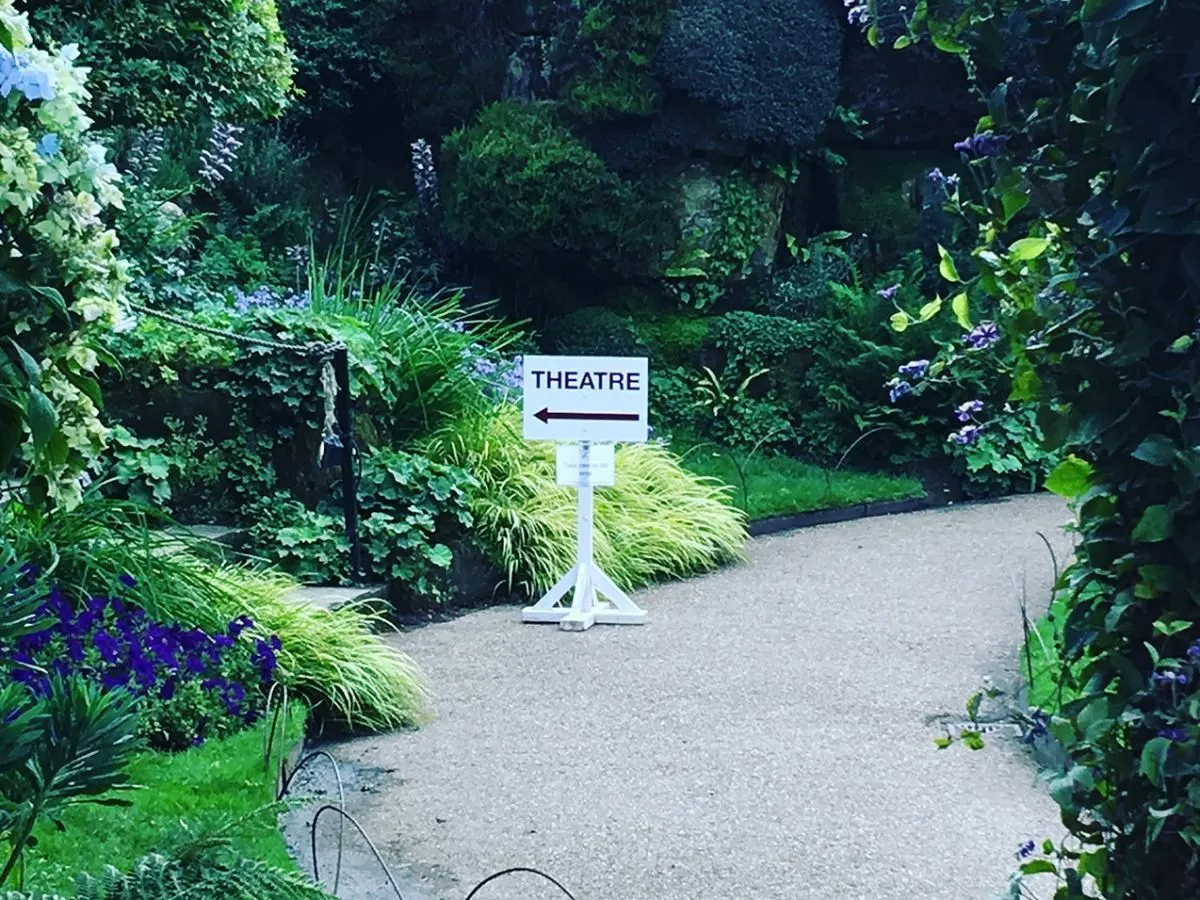 A directional sign reading "THEATRE" with an arrow pointing left stands on a garden path surrounded by lush green foliage and purple flowers.