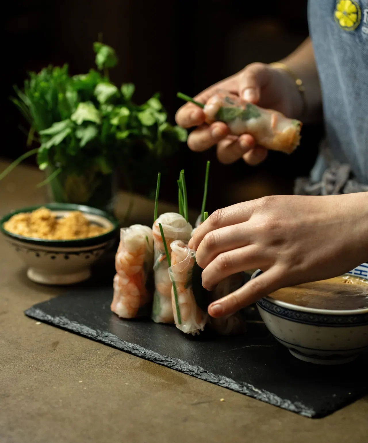 Two people's hands preparing spring rolls with fresh herbs, served with dipping sauce at a cookery school.
