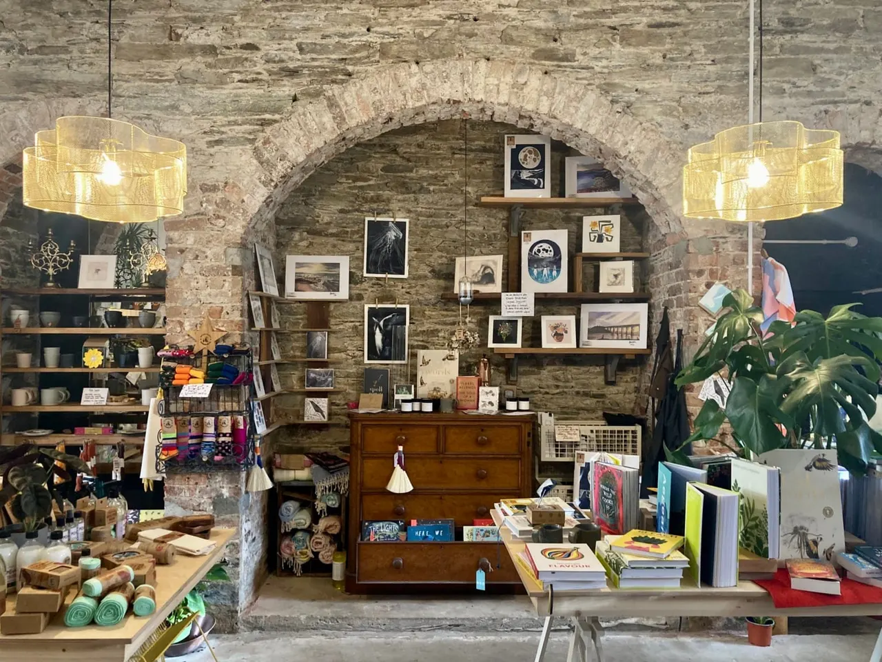 A stone-vaulted shop interior displaying artwork, books, plants and crafts on shelves and a wooden chest.