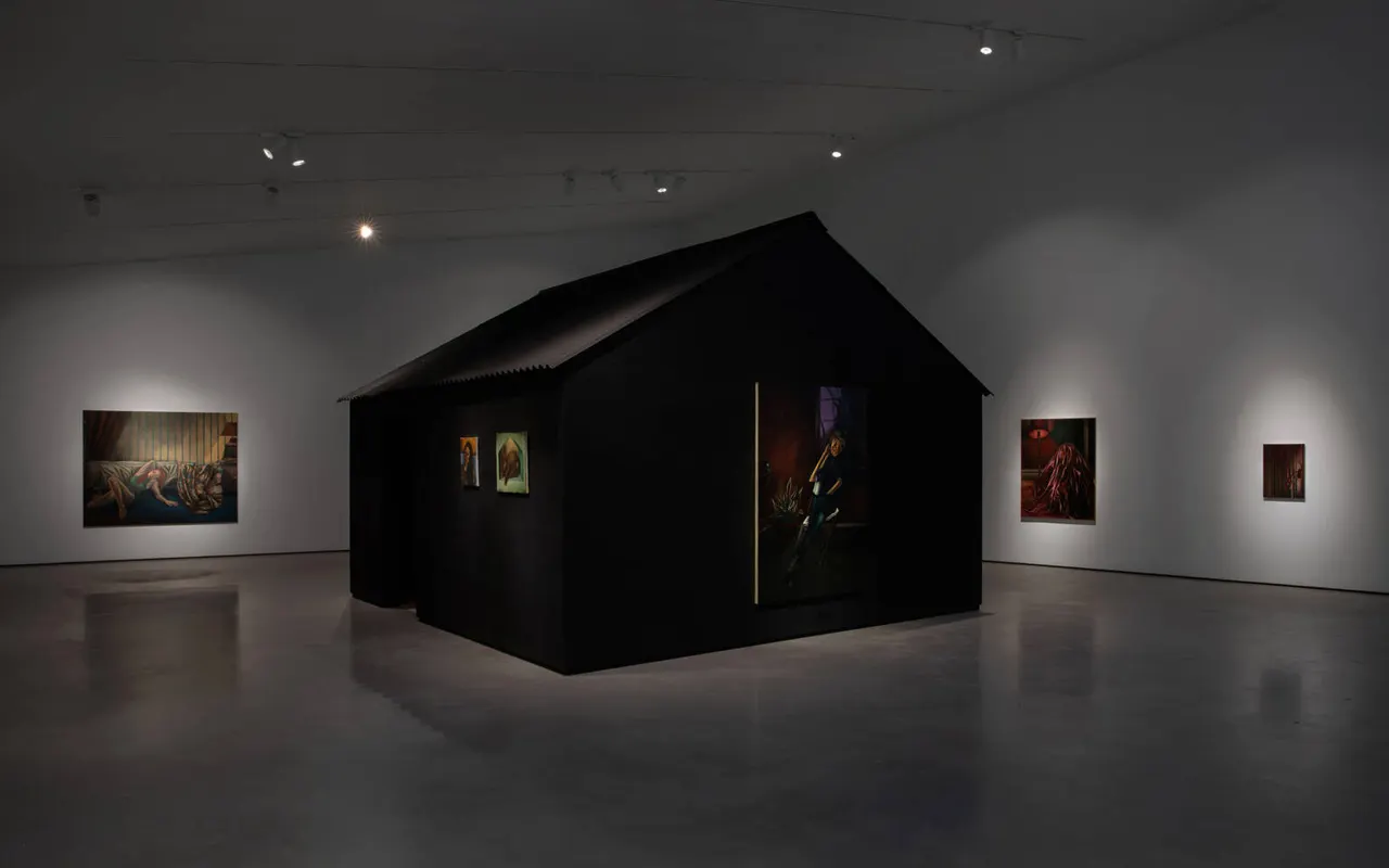 Modern art gallery interior with a large black structure in the centre displaying artwork, surrounded by illuminated paintings on white walls.