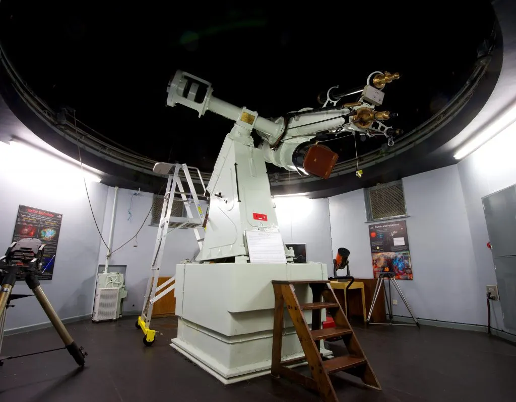 Large white telescope mounted inside a domed planetarium with visitor controls and information panels visible.