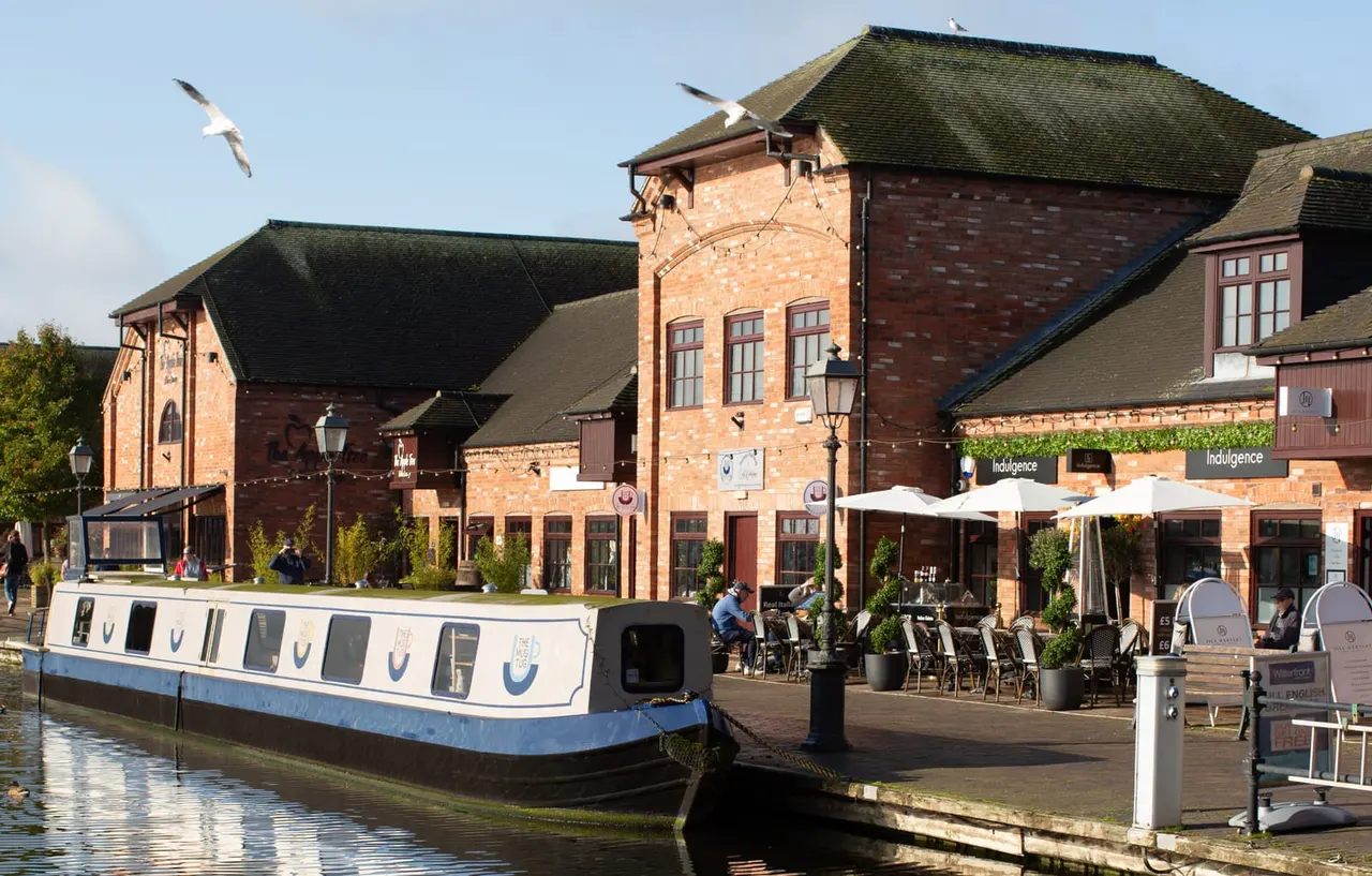 A narrowboat moored at a canalside venue with brick buildings, outdoor seating under parasols, and two large birds flying overhead.
